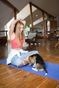 A young woman sits on a yoga mat indoors, with a beagle puppy nearby, in a serene home setting.