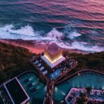 Aerial view of Savaya Bali in Uluwatu at sunset, showing the illuminated Cube stage, clifftop pools and crowd above the Indian Ocean