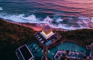 Aerial view of Savaya Bali in Uluwatu at sunset, showing the illuminated Cube stage, clifftop pools and crowd above the Indian Ocean